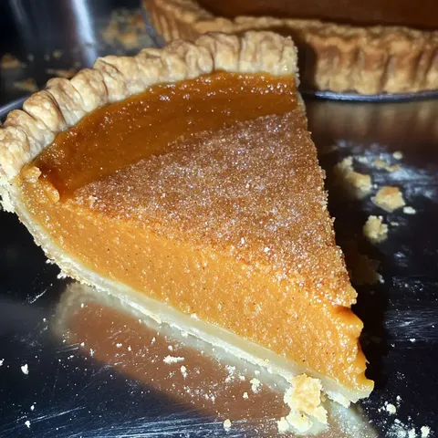 Ein Stück Kürbiskuchen mit knusprigem Teig und glatter Füllung auf einer glänzenden Oberfläche.