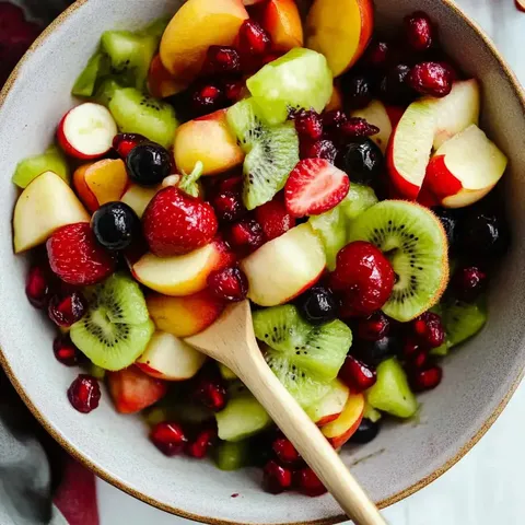 Eine farbenfrohe Obst-Mischung mit Apfel-, Erdbeer-, Kiwi- und Melonenstückchen, kombiniert mit Himbeeren und Granatapfelkernen, in einer Schüssel mit Holzlöffel serviert.