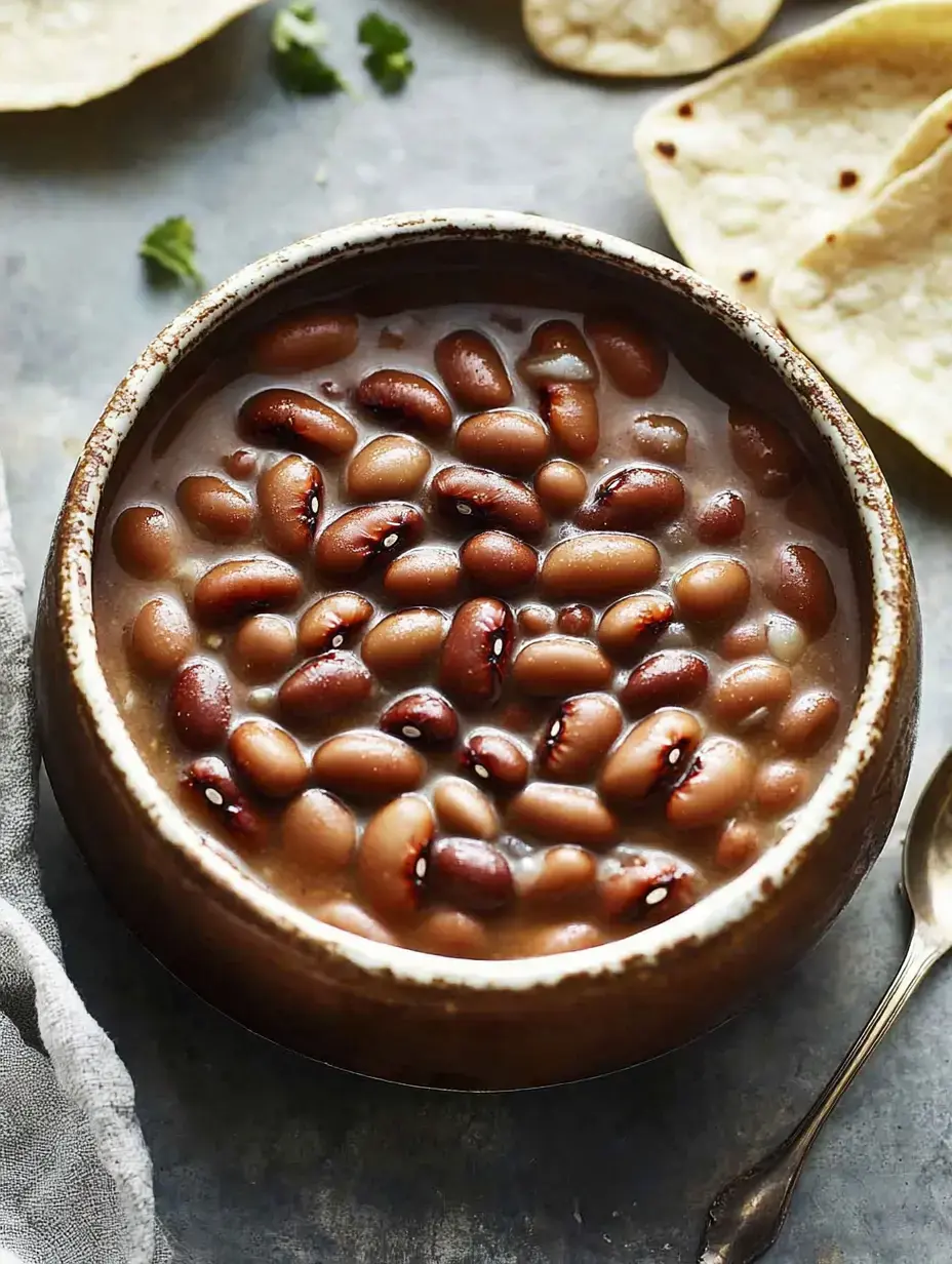 Eine Schüssel Bohnen im Sud steht neben frischen Tortillas und Koriander auf rustikalem Tisch.