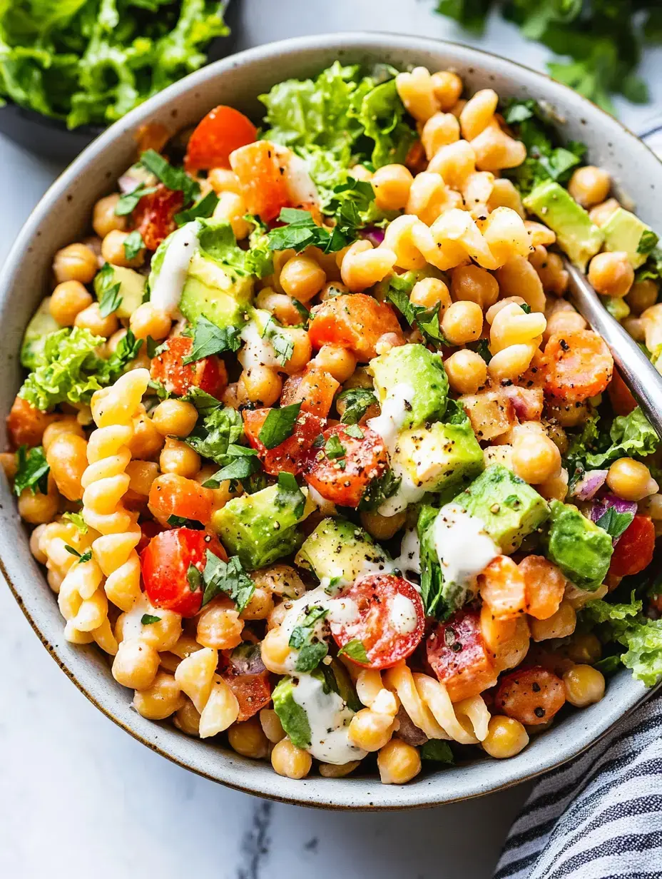 Eine bunte Schale mit Nudelsalat, Kichererbsen, Cherrytomaten, Avocado, Blattsalat und cremigem Dressing, obenauf frische Kräuter.