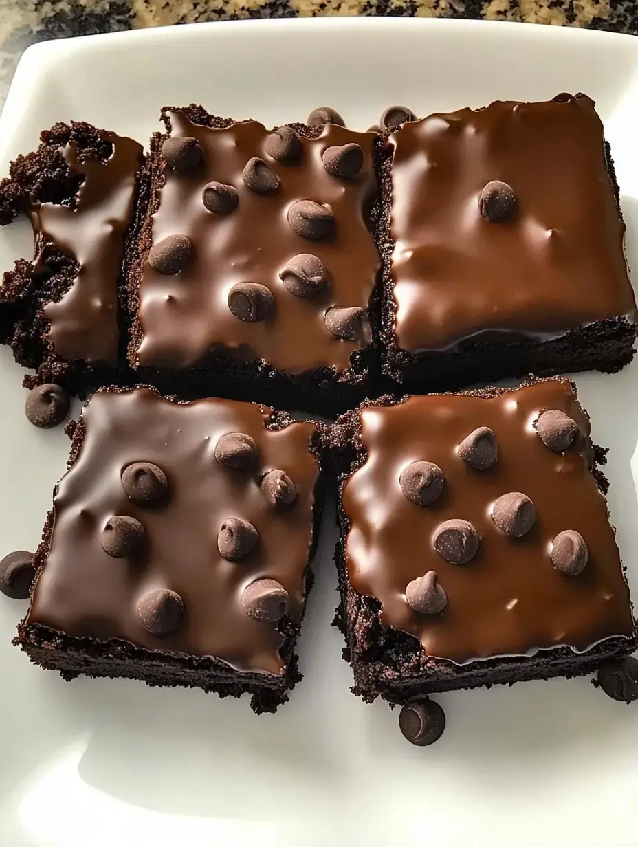 Nahaufnahme von vier Brownies mit glänzendem Schokoguss und Schokostücken auf einem weißen Teller.