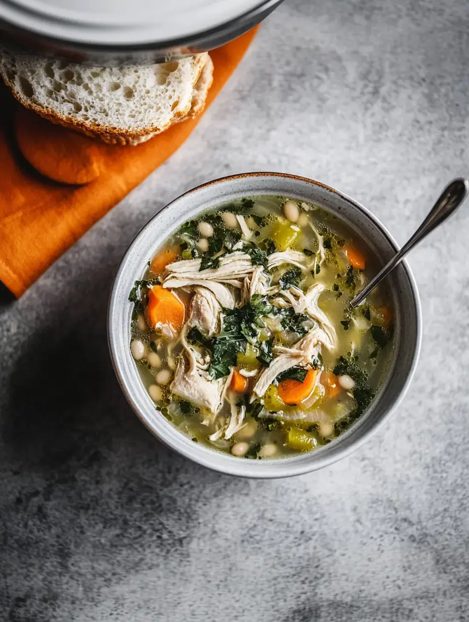 Eine Schüssel Hähnchensuppe mit zerrupftem Huhn, Karotten, Blattgemüse und weißen Bohnen, dazu ein Stück Brot auf grauer Struktur.