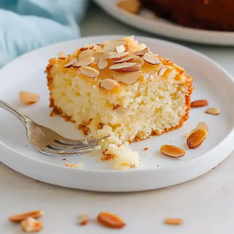 Lockerer Kuchen mit Mandelschicht auf einem Teller, dekoriert mit Mandelblättchen, daneben liegt eine Gabel.