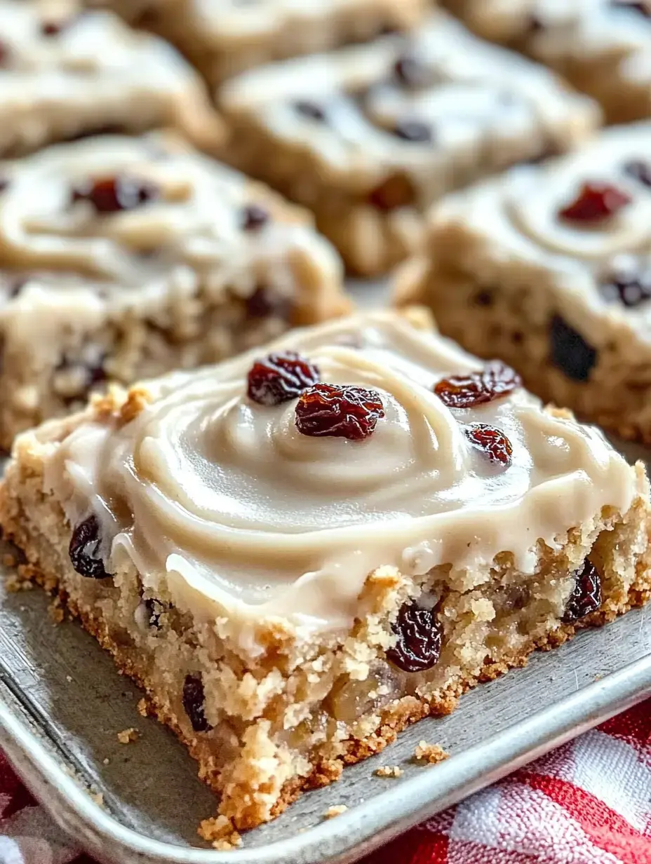 Nahaufnahme eines saftigen Kuchenstücks mit glattem Frosting und Rosinen oben drauf, im Hintergrund weitere Stücke auf einem Tablett.