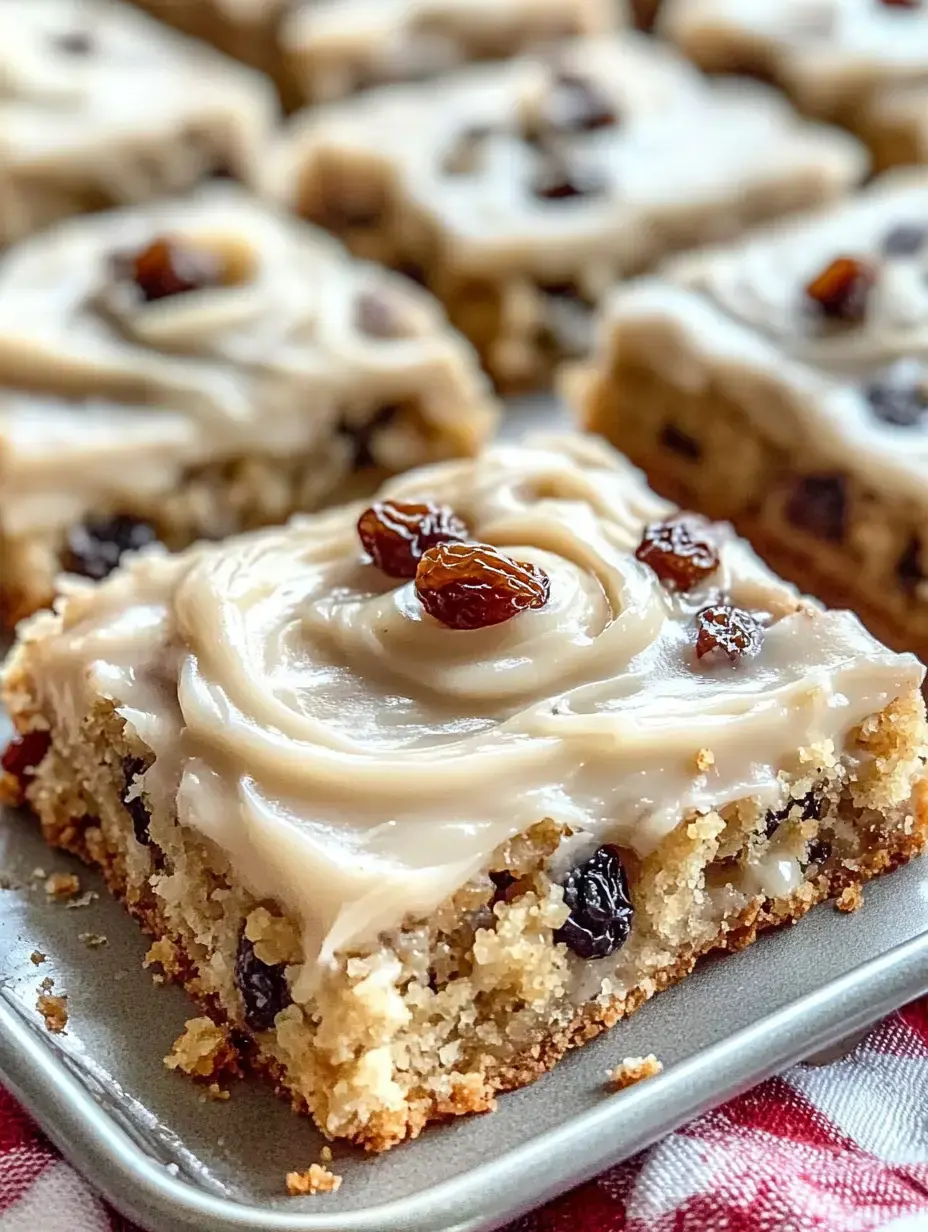Nahaufnahme eines quadratischen, gefrosteten Kuchenstücks mit Rosinen auf einem Teller, weitere Stücke im Hintergrund.